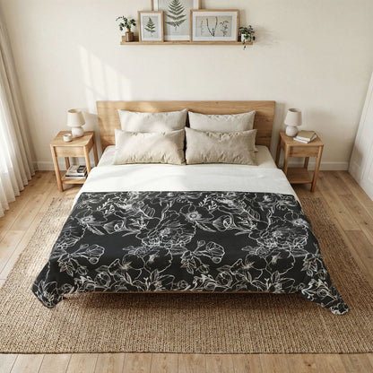 A neatly made bed in a bright, minimalist bedroom, featuring a black comforter with a white floral line-art pattern over white sheets and beige pillows.